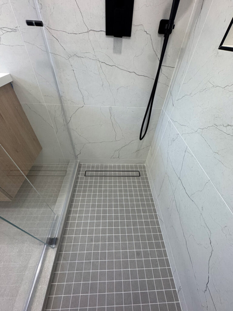 San Jose bathroom remodel looking down into the shower, the gray square mosaic base and its black linear drain framed by porcelain walls with soft veins and the frameless glass return.