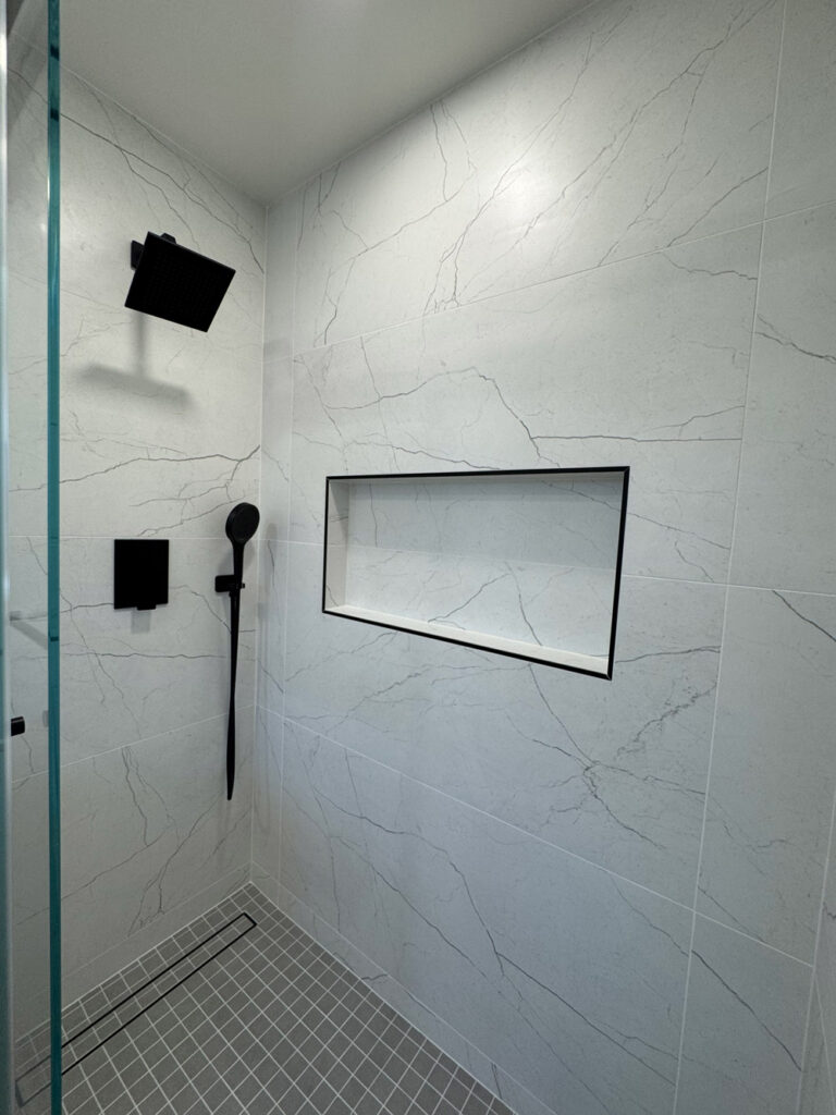 San Jose bathroom remodel shower corner with a black rain head and handheld on the left wall and a long shampoo niche trimmed in black Schluter profile recessed into the porcelain with soft veins opposite, above gray mosaic flooring.