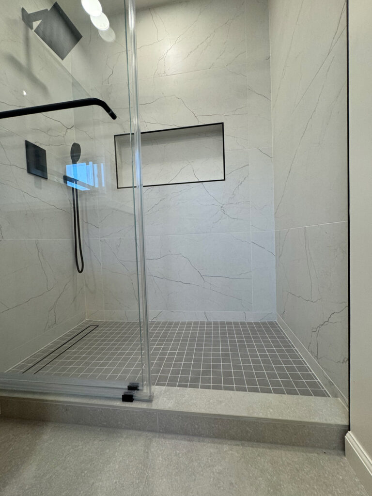 San Jose bathroom remodel shower base seen from the doorway, with a black grab bar, shampoo niche and rain head above the gray mosaic floor, stepping down to concrete-look tile outside the enclosure.