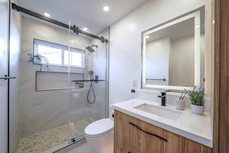 San Jose bathroom remodel wide view with the sliding glass shower on its black rail, a clerestory window trimmed in black Schluter profile, a pebble mosaic shower floor, and a walnut vanity with a lit mirror beyond the toilet.