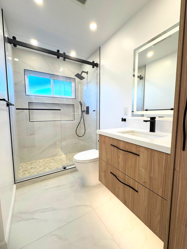 San Jose bathroom remodel looking into the shower through the sliding glass door, with an obscured-glass window glowing blue, a shampoo niche trimmed in black Schluter profile, black rain and handheld showers, and a pebble mosaic floor.