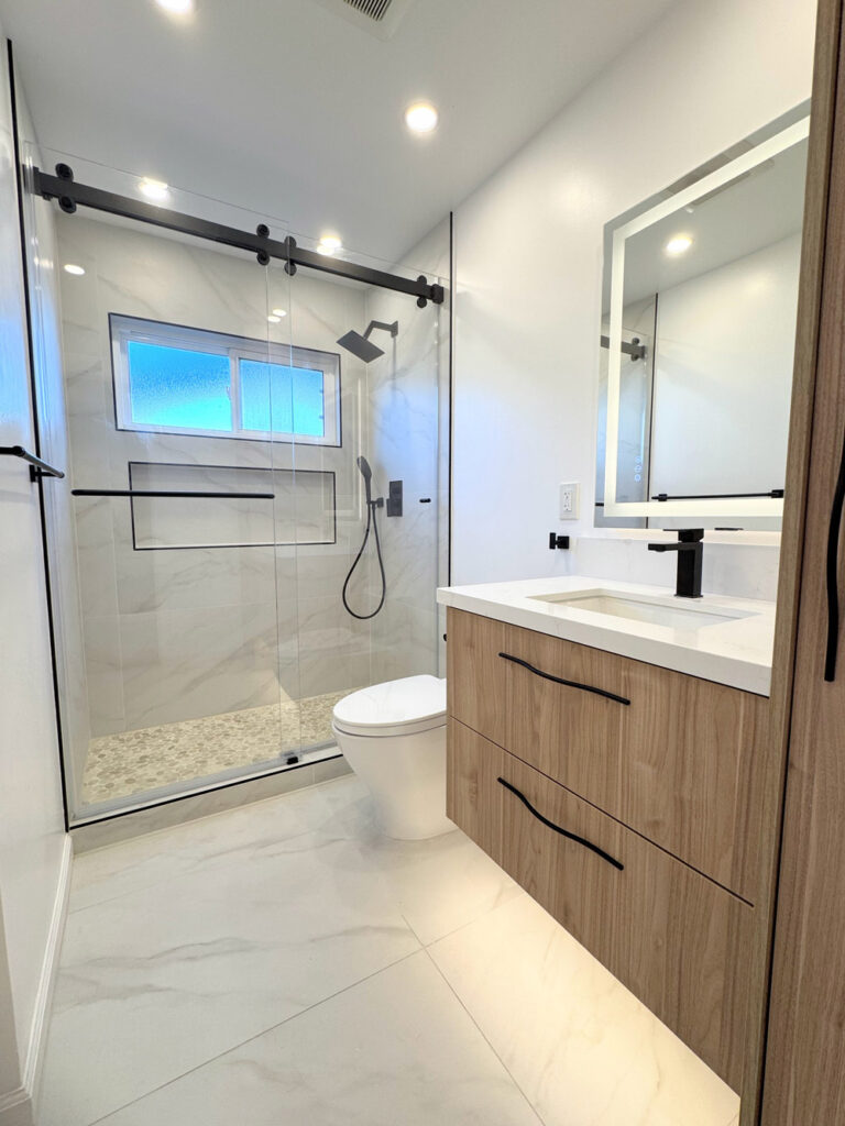 San Jose bathroom remodel with the sliding shower enclosure filling the left of the frame, its black roller rail and towel bar set against porcelain with soft veins, and the lit mirror and walnut vanity at right.
