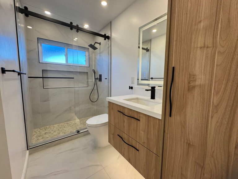 San Jose bathroom remodel showing the tall walnut linen cabinet at right, the floating vanity with black faucet and backlit mirror, and the glass shower with its window and pebble floor at left.